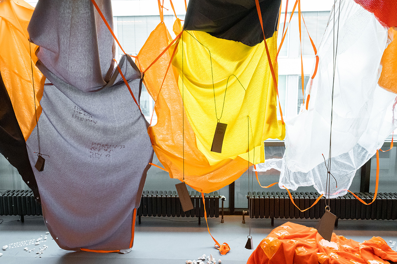 A wide angle photo of the Pop Up Disabled Data Center shows three large and colorful sails made from differently textured fabrics in front of large windows. The left sail is a soft violet sewn together with smaller orange and grey fabrics and has braille embroidered. The middle sail falls in round curvings and is bright yellow, orange and grey. The sail on the right is made from bubblewrap that shimmers in opacities and hovers above an orange blanket placed below. The sails are connected via orange straps that weave in and out of embedded metal gromets, and that are also holding them via attachments on the ceiling. From these sails, grey booklets containing the data sets dangle downwards on long grey strings that permeate the fabrics. Below, small clay figures are scattered across on the floor.