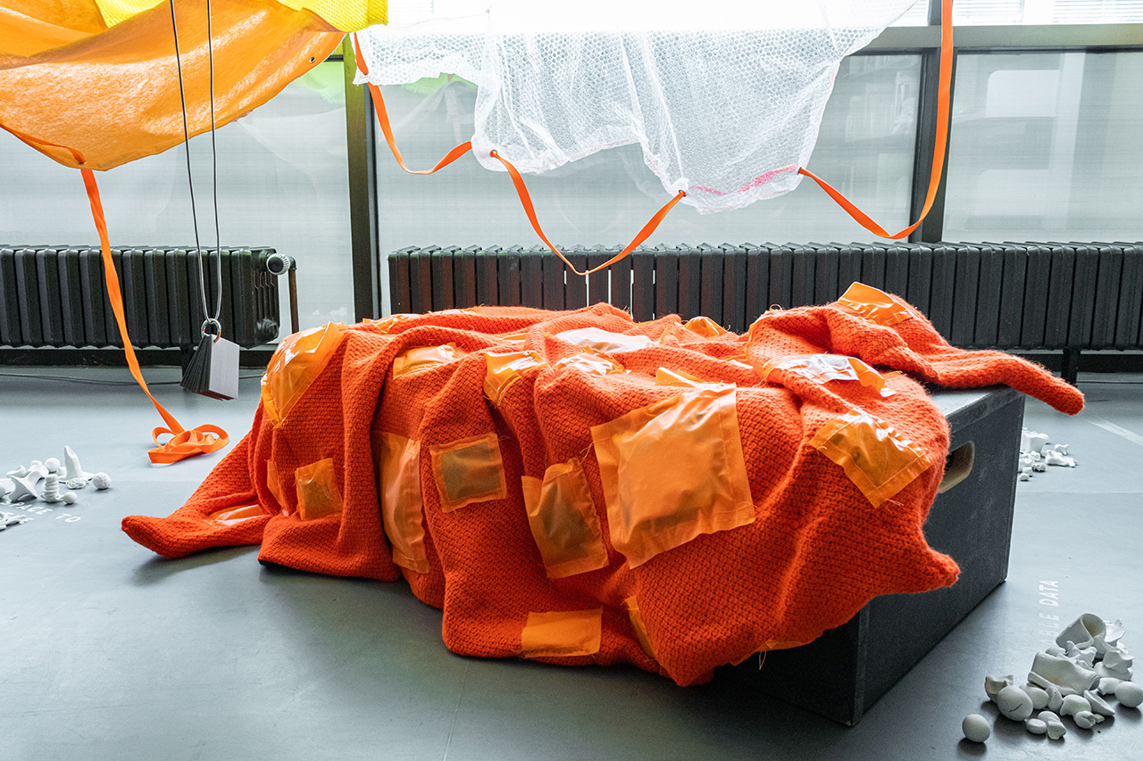 datasetofweight_01.jpg In the middle of this photo, an orange blanket floats, folds and rests across a dark grey bench. The blanket is made from a warm knitted textile, while the pockets are shiny and look smooth to the touch. From viewing alone, the contents of the pockets cannot be determined, but it is clear that the materials are different. Around the blanket, there is floor space with small white clay figures arranged around vinyl fonts. Above, there are two fabric sails: One is an orange textile, one is a fabric made from bubblewrap. On the left, a booklet is dangling down from above so that it can be picket up and read when sitting down and using the blanket.