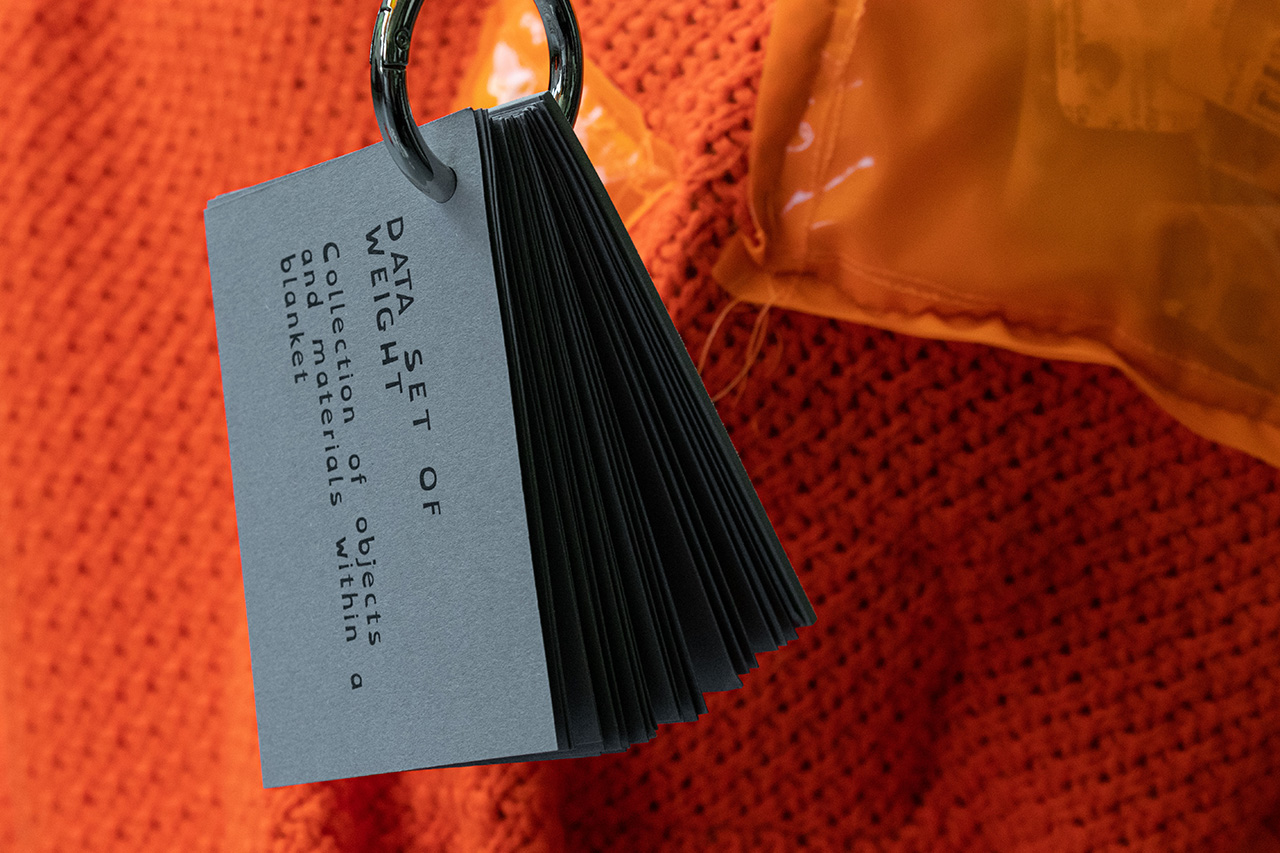 A grey booklet with the title "Data Set of Weight. Collecting of objects and materials within a blanket" held together by a large metal ring is dangling in front of a blanket made from knitted orange textile onto which a glossy plastic-like pocket is sewn on the right.