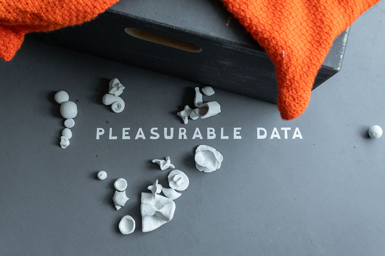On a grey floor, the letters "Pleasurable Data" appear in vinyl. Around them, small clay sculptures are arranged in rows and heaps, forming various proximities and relations. From the top of the photo, a grey box with an orange woolen blanket enters the image.