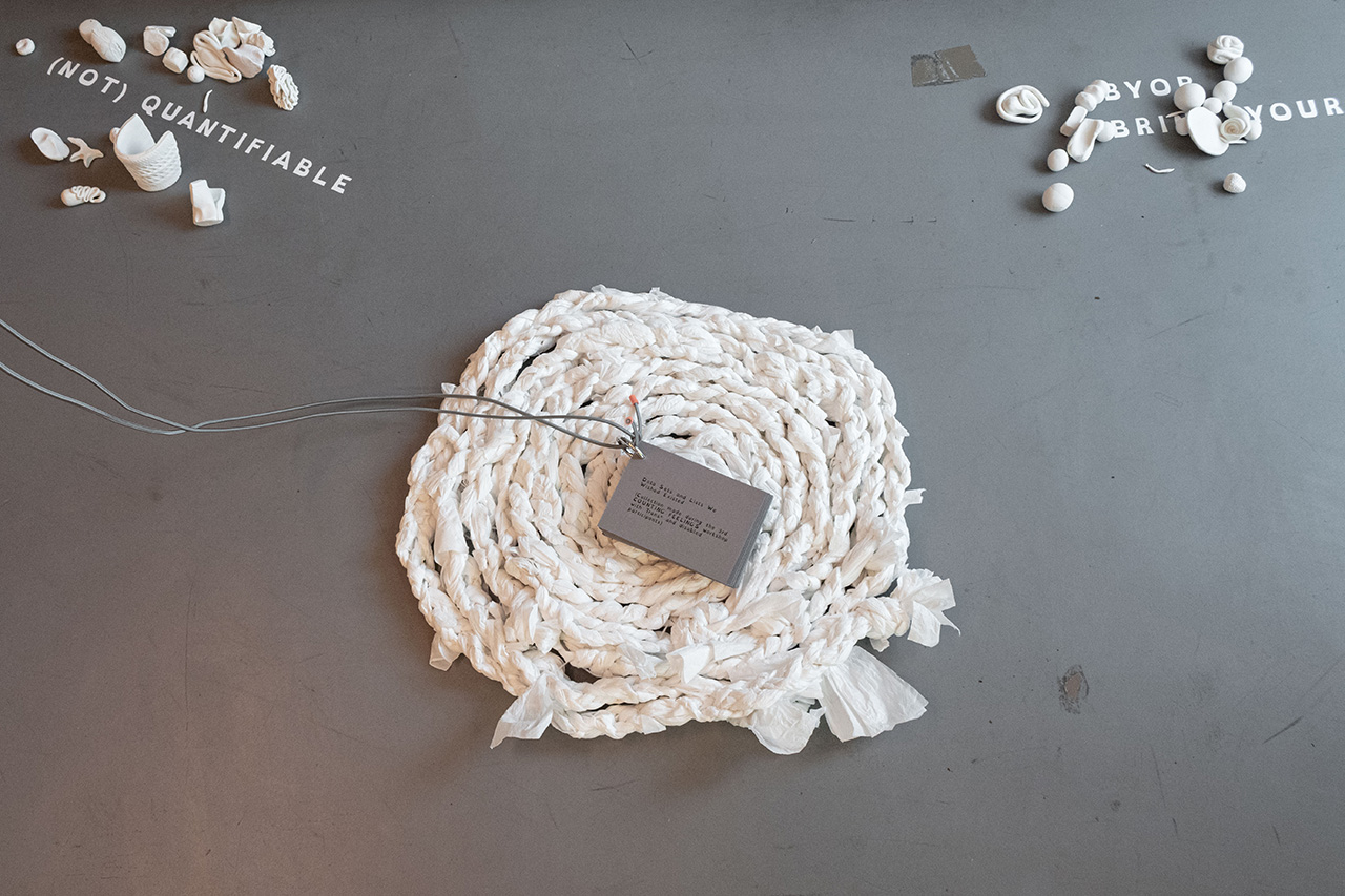 Photographed from above, this image shows a flat pillow made from braided plastic ropes, on the center of which a grey booklet connected to a string rests. Two other elements on the grey floor are two statements printed in vinyl that read "(not) quantifiable" and "BYOB" with some more letters covered by small clay figures. More clay figures are arranged around these statements.